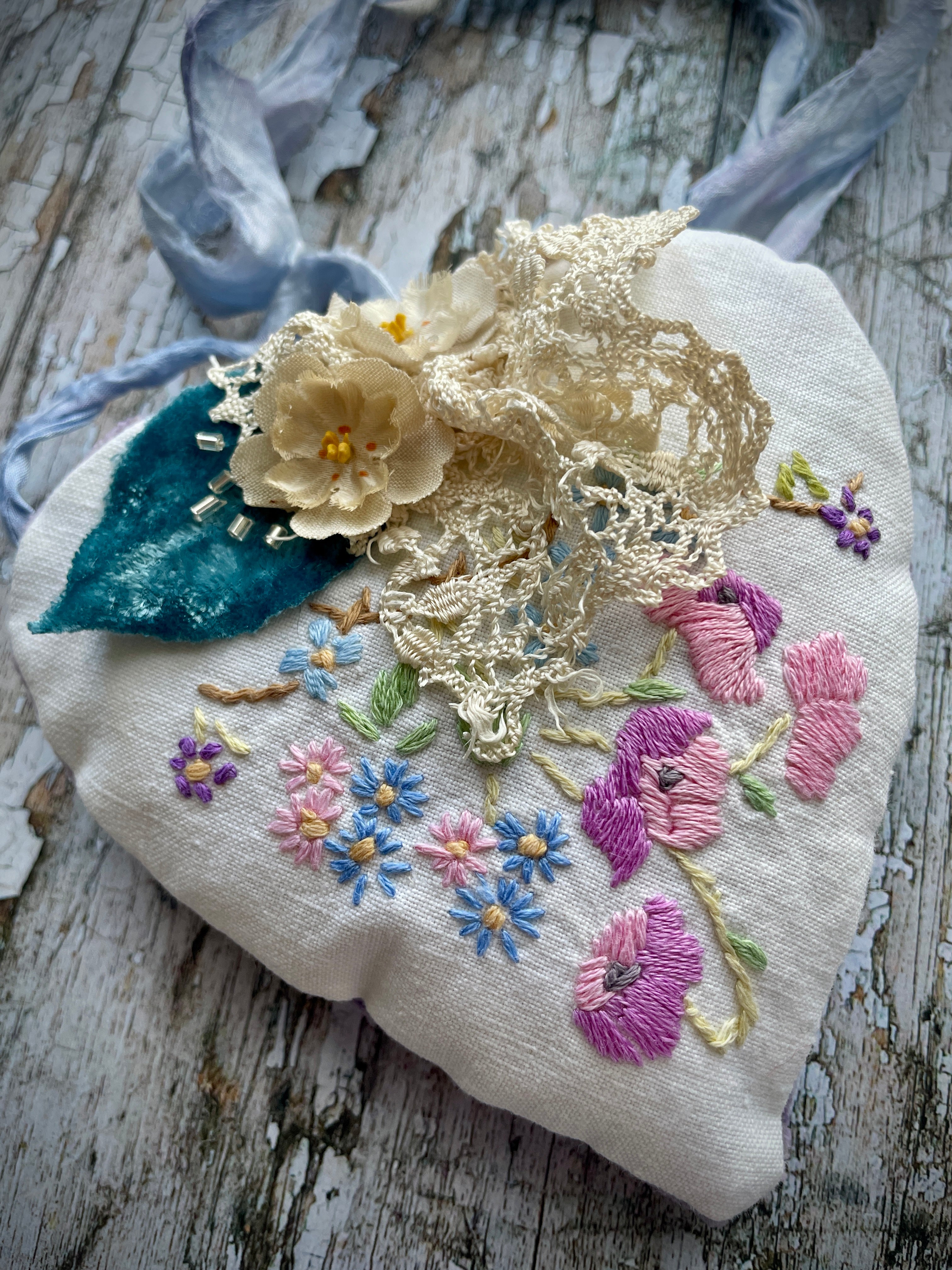 A beautiful one of a kind heart... made by hand stitching together vintage embroidered linen of pink, mauve, lavender and blue flowers, with vintage cream lace, linen flowers and a blue velvet leaf embellished with small silver bugle beads. The back is made of pale lavender velvet and the heart hangs with a loop of blue raw edged sari silk to wear around the neck or hang on the wall as a decoration. The inside of the heart has been gently filled with non-allergenic filling conforming to BS + CE  standards.