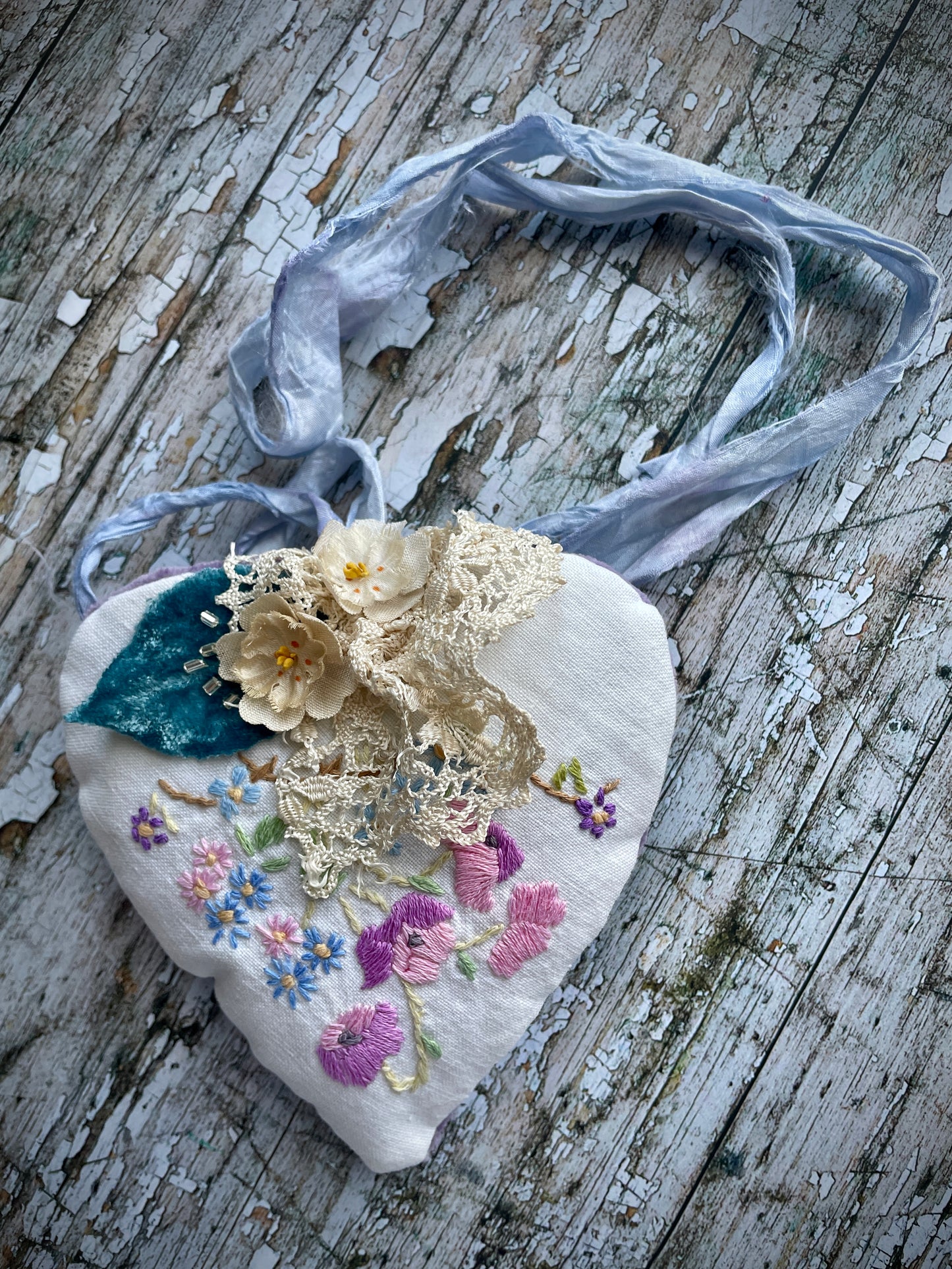 A beautiful one of a kind heart... made by hand stitching together vintage embroidered linen of pink, mauve, lavender and blue flowers, with vintage cream lace, linen flowers and a blue velvet leaf embellished with small silver bugle beads. The back is made of pale lavender velvet and the heart hangs with a loop of blue raw edged sari silk to wear around the neck or hang on the wall as a decoration. The inside of the heart has been gently filled with non-allergenic filling conforming to BS + CE  standards.