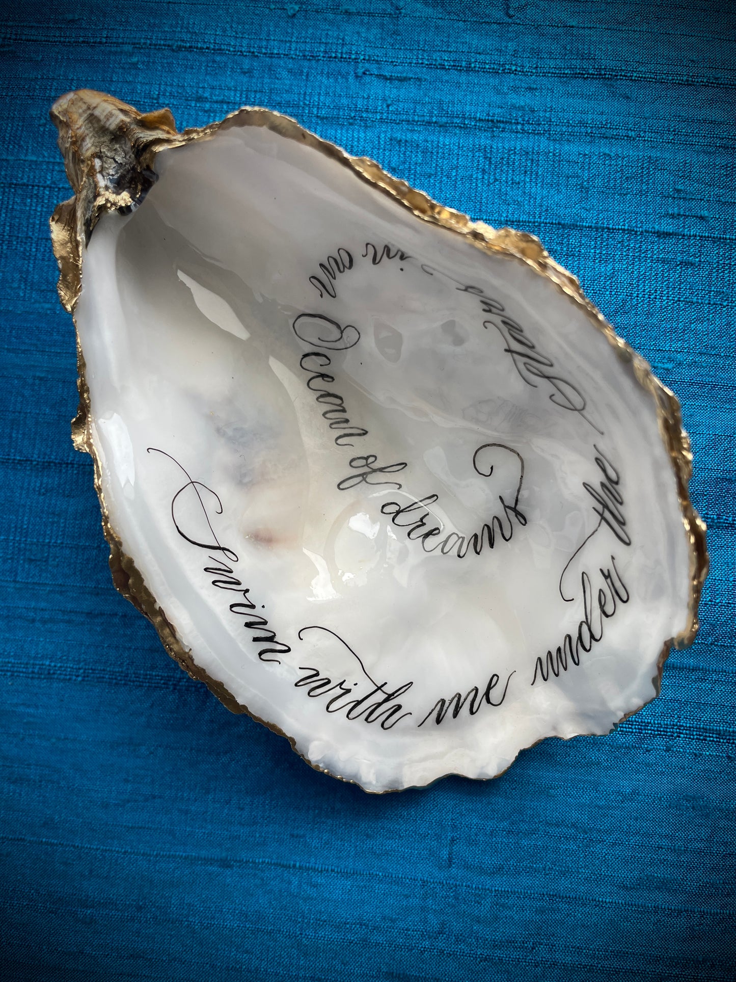 Cornish Oyster Shell Calligraphy

Made by: Julia Brooke - a word in your shell
'Swim with me under the stars in an ocean of dreams'
The inside of this Cornish oyster shell has been beautifully hand written with a romantic message, edged with gold leaf and the back lightly burnished with gold wax... perfect as a romantic gift.
The inside has been protected with clear uv stable resin making it smudge proof and moisture resistant, but not for aquatic display.
Size approx: 11.5 cm x 7 cm / 4+1/2" x 2+3/4"