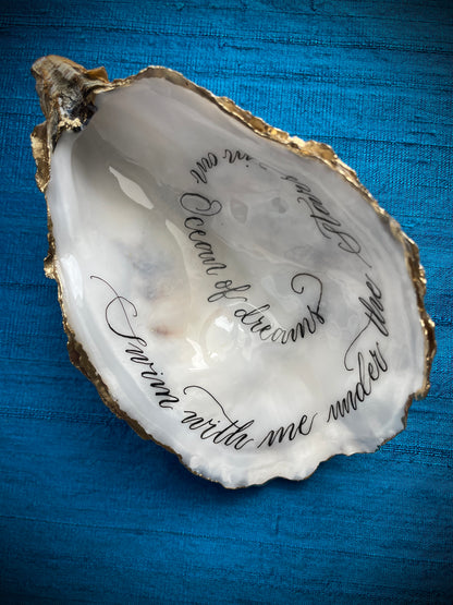 Cornish Oyster Shell Calligraphy

Made by: Julia Brooke - a word in your shell
'Swim with me under the stars in an ocean of dreams'
The inside of this Cornish oyster shell has been beautifully hand written with a romantic message, edged with gold leaf and the back lightly burnished with gold wax... perfect as a romantic gift.
The inside has been protected with clear uv stable resin making it smudge proof and moisture resistant, but not for aquatic display.
Size approx: 11.5 cm x 7 cm / 4+1/2" x 2+3/4"