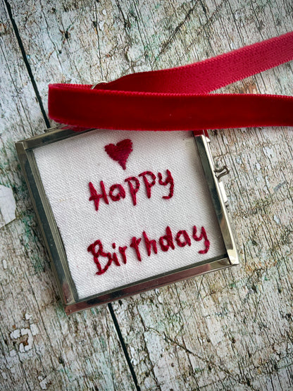 Romantic Happy Birthday Keepsake

The words 'Happy Birthday' and a heart have been beautifully hand embroidered onto a piece of vintage French linen with deep red silk thread. It has been backed with a vintage floral cotton fabric, then placed inside a sealed small glass and silver coloured metal frame with a deep red velvet hanging loop. Perfect as a small birthday keepsake gift.
Frame size approximately: 5 cm / 2" square.
Velvet loop length approximately: 15cm / 6" long.