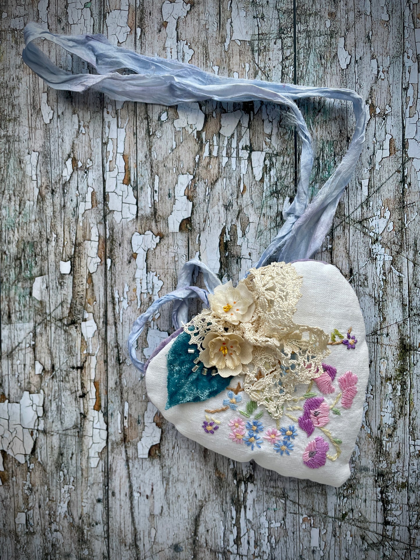 A beautiful one of a kind heart... made by hand stitching together vintage embroidered linen of pink, mauve, lavender and blue flowers, with vintage cream lace, linen flowers and a blue velvet leaf embellished with small silver bugle beads. The back is made of pale lavender velvet and the heart hangs with a loop of blue raw edged sari silk to wear around the neck or hang on the wall as a decoration. The inside of the heart has been gently filled with non-allergenic filling conforming to BS + CE  standards.